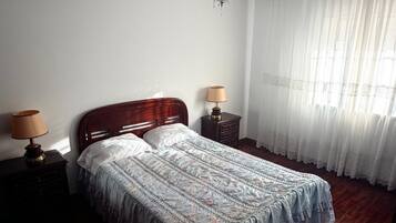 A bedroom with a bed, two bedside tables with lamps, a chandelier, and a window with sheer curtains.