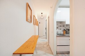 Un pasillo estrecho con un banco de madera, paredes blancas y suelo de baldosas que conduce a una zona de cocina con armarios blancos y un lavamanos.