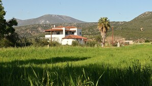 A two-story building with a red-tiled roof, surrounded by a grassy field and a mountain in the background. - Apartment 'The Rustic Archontiko' with Sea View (Kranidi)