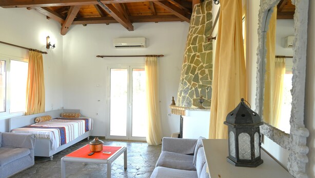 A cozy room with a stone wall, a bed with a patterned blanket, a small table with a pot and remote, and a lamp. - Apartment 'The Rustic Archontiko' with Sea View (Kranidi)