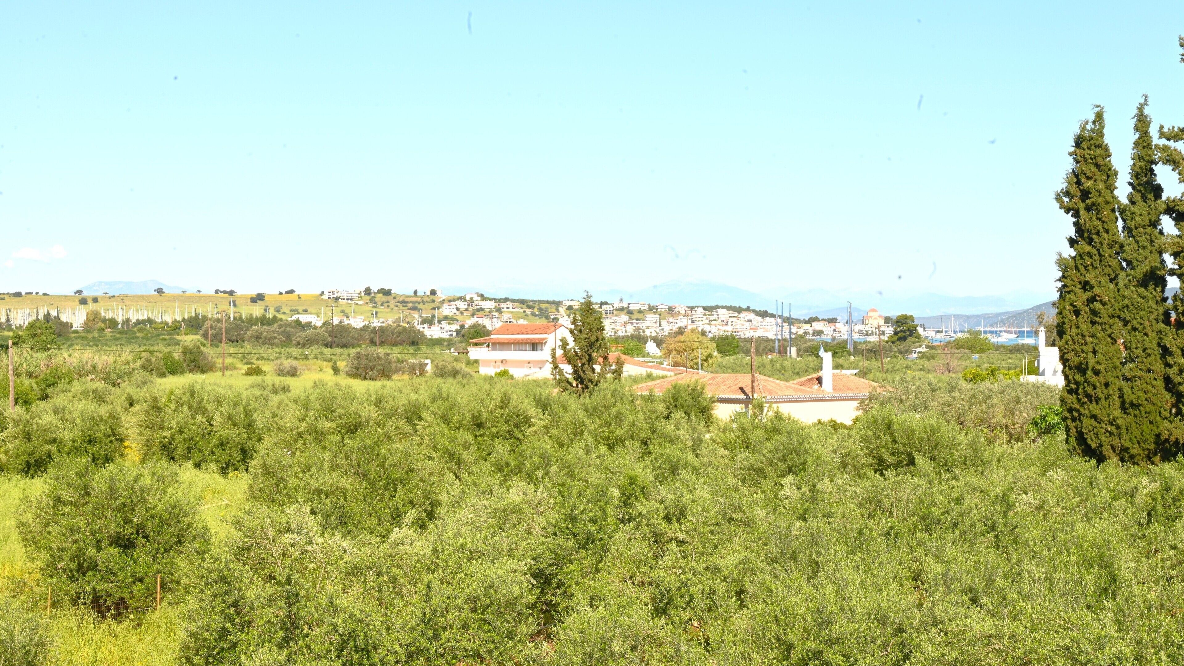 Paisatge rural amb un edifici envoltat de vegetació, un poble a la distància i muntanyes al fons.