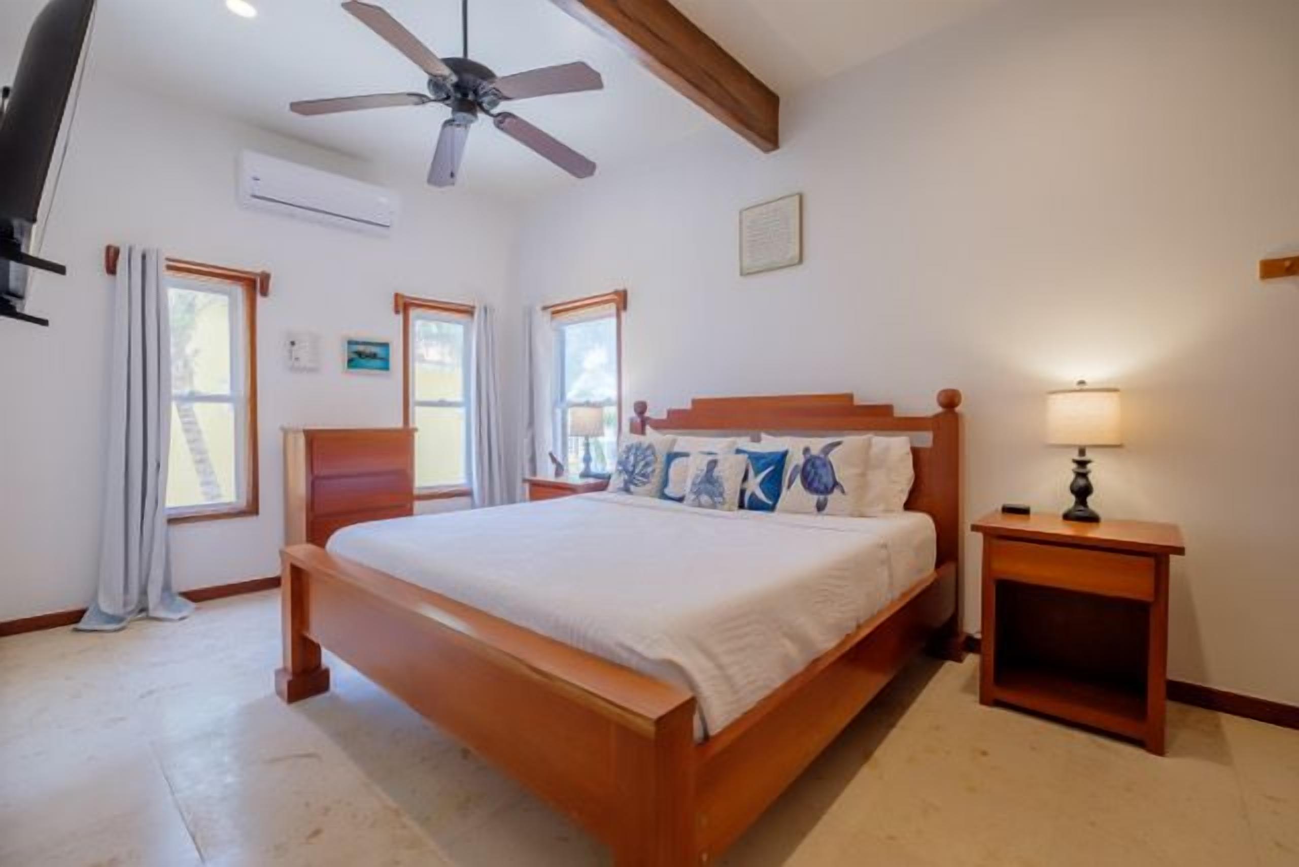 Un dormitorio con una cama de madera, un ventilador de techo, una mesita de noche con lámpara y una ventana con cortinas.