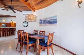 Un coin repas avec une table et des chaises en bois, un ventilateur de plafond, une horloge murale, et une cuisine en arrière-plan.
