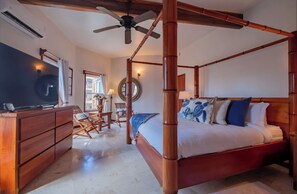 Un lit à baldaquin avec un ventilateur de plafond, un téléviseur posé sur une commode en bois, et un coin salon avec des fauteuils et une petite table.