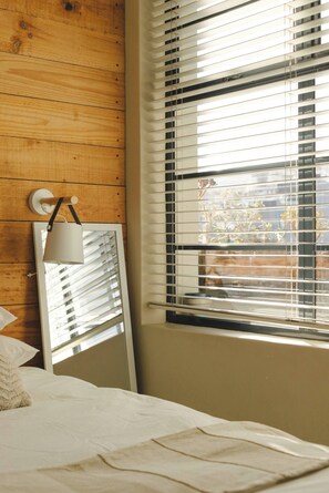 A bedroom with a wooden wall, a bed with white bedding, a bedside lamp, and a window with blinds. - Central Cape Town Apt With Views (Cape Town)