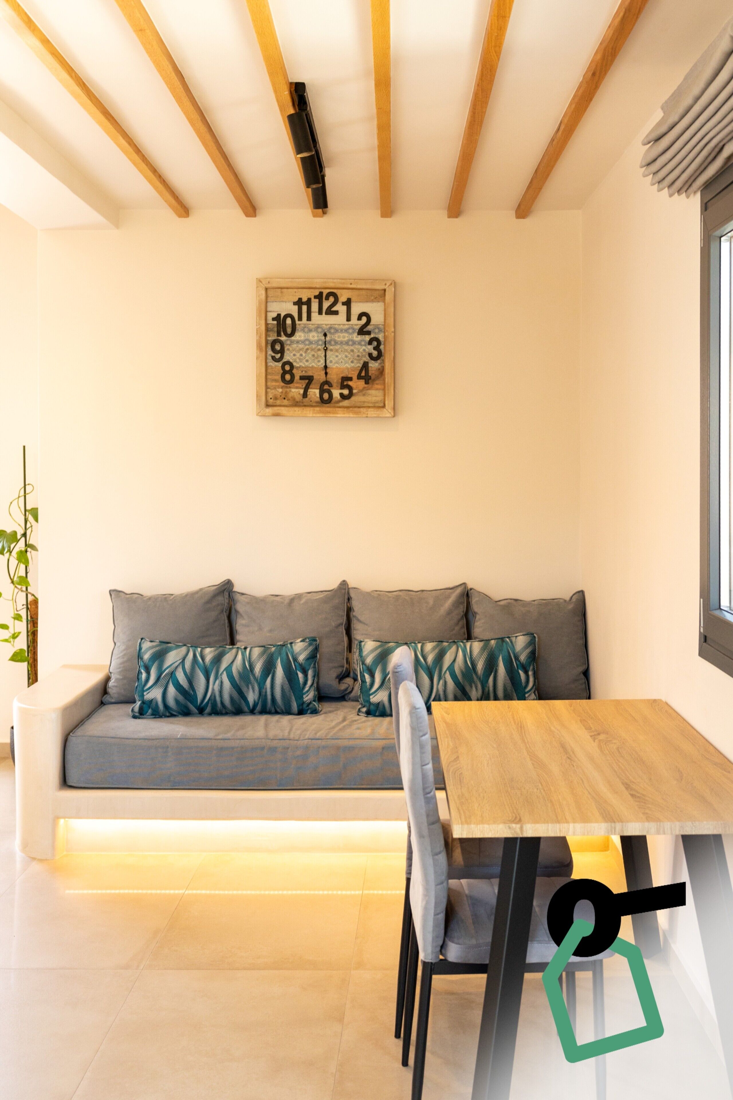 A modern living room with a wooden ceiling, a sofa with patterned cushions, a wooden dining table, and a wall clock.