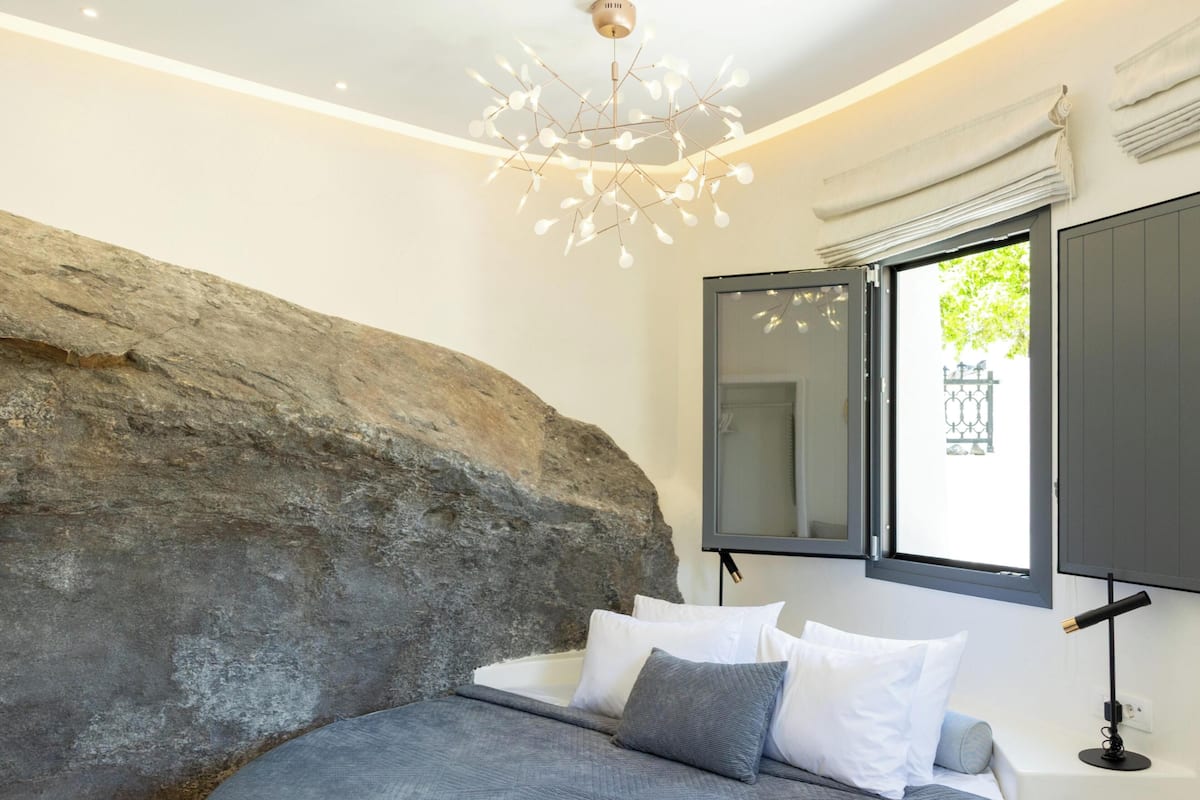 A bedroom with a large rock wall, a bed with grey bedding, a modern chandelier, a window with curtains, and a bedside table with a lamp.