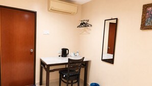 A small room with a wooden door, a table with a chair, a black trash bin, a blue bag, a black kettle, a white cup, a black hanger, and a wall-mounted air conditioner. - The Ardens Hotel - Pandan Johor Bahru (Johor Bahru)