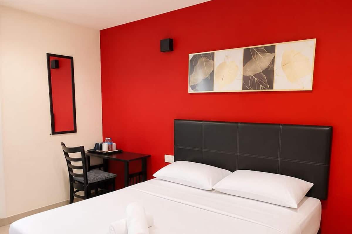 Quarto de hotel com uma cama grande, uma escrivaninha com cadeira, uma parede de destaque vermelha e uma luminária no teto.