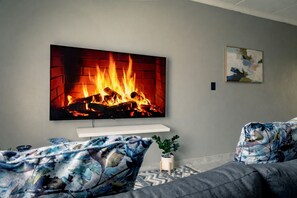 Ein Wohnzimmer mit einem großen Fernseher, der einen Kamin zeigt, einem Sofa, einer kleinen Pflanze und einem Bild an der Wand.