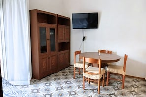Una habitación con un armario de madera, una mesa redonda con sillas, un televisor montado en la pared y baldosas estampadas en el suelo.
