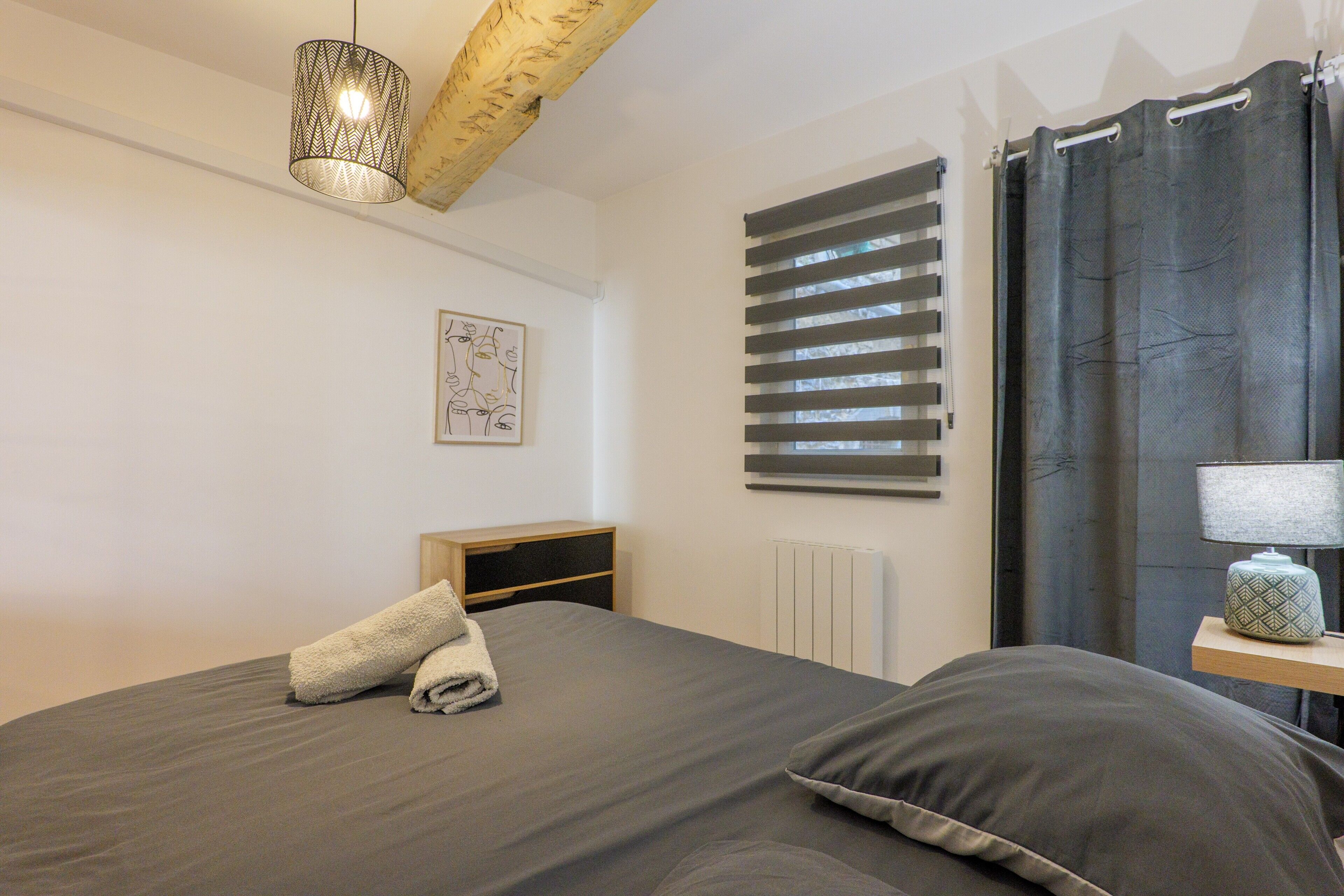 A bedroom with a bed, a bedside table with a lamp, a radiator, and a window with blinds.