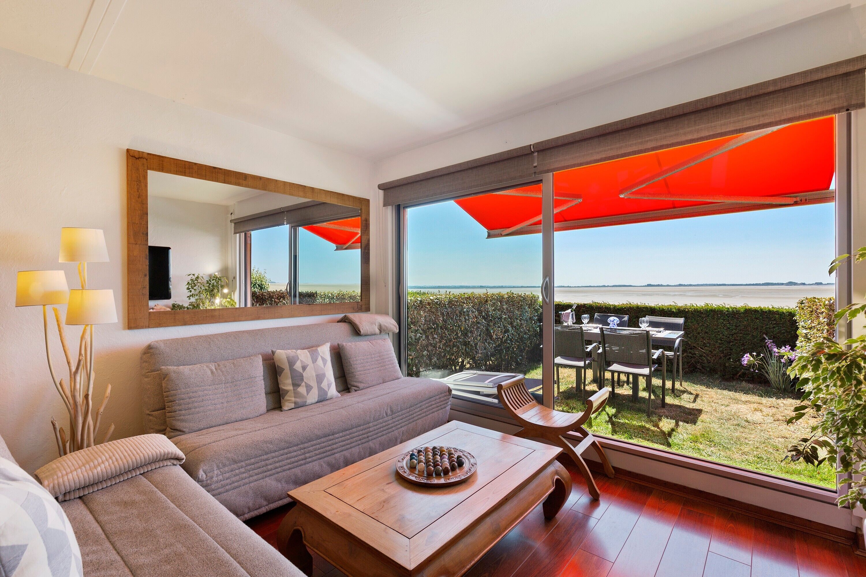 A modern living room with a large window, a sofa, a coffee table, and a dining area with outdoor seating.