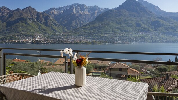 Ein Balkon mit Tisch und Blumen, mit Blick auf einen See und Berge.