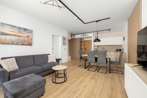 Ein modernes Wohnzimmer mit Sofa, Hocker und rundem Tisch. Es gibt einen Essbereich mit Stühlen und einem Tisch. Im Hintergrund ist eine Küche mit Schränken und einer Arbeitsplatte zu sehen.