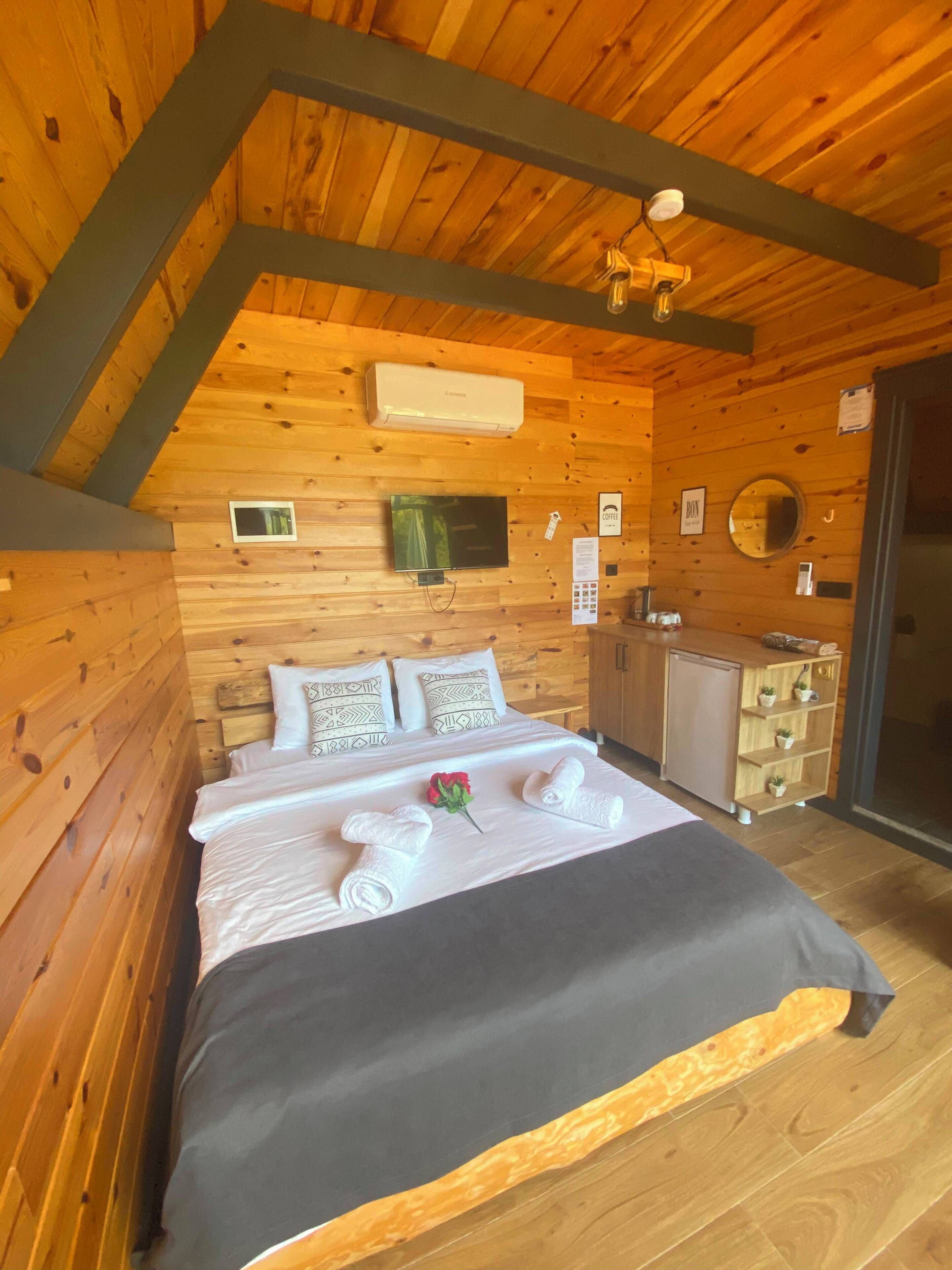 A wooden cabin bedroom with a bed, pillows, a small TV, an air conditioner, a mirror, and a wooden cabinet.