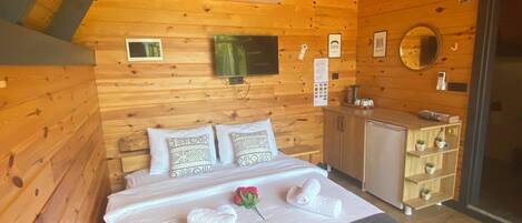 A wooden cabin bedroom with a bed, pillows, a small TV, an air conditioner, a mirror, and a wooden cabinet.