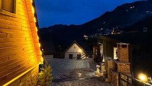 A stone-paved pathway leading to a wooden building with a sloped roof, surrounded by mountains.