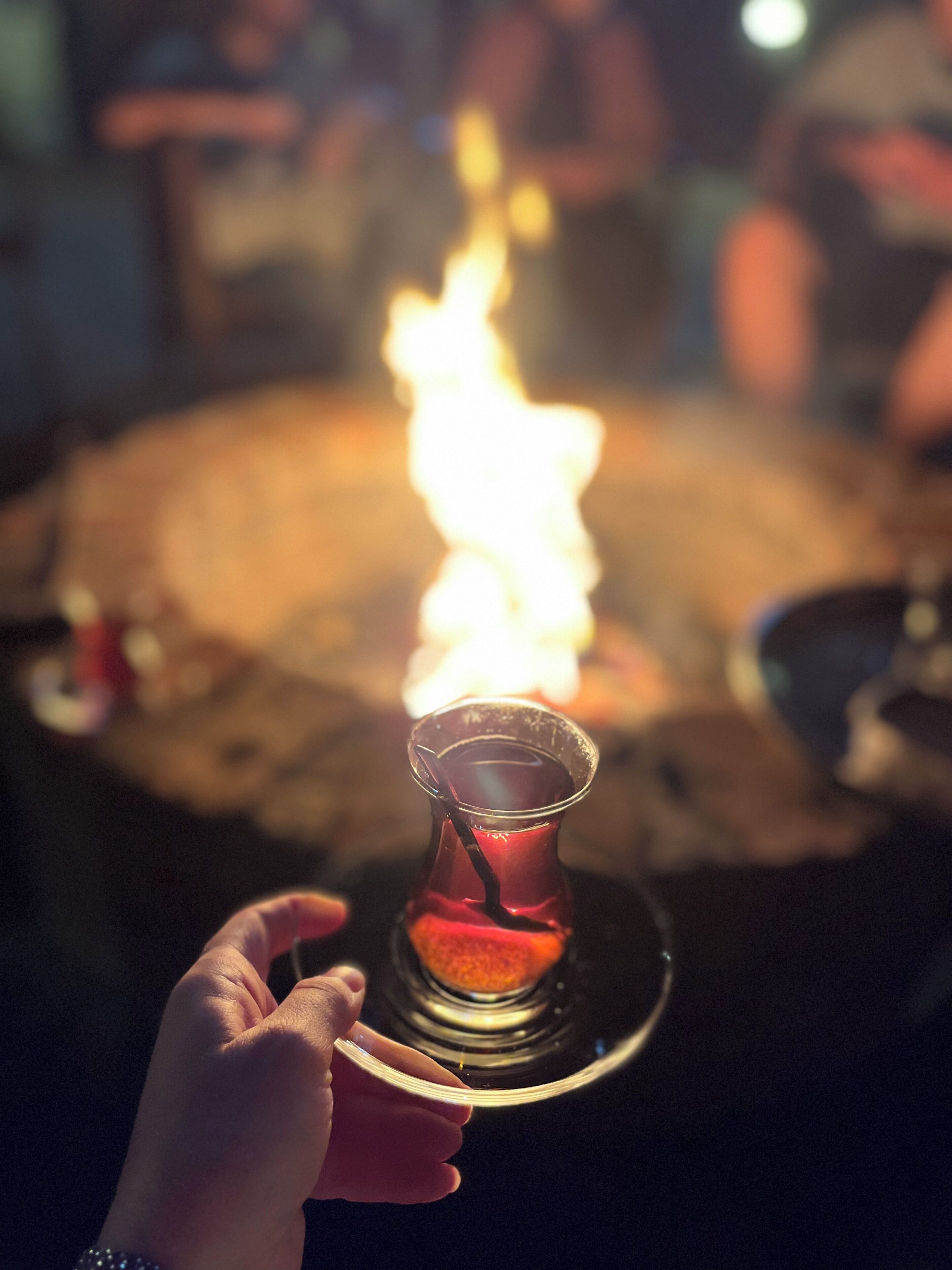 A hand holding a glass with a drink, a fire in the background, and a blurred crowd.