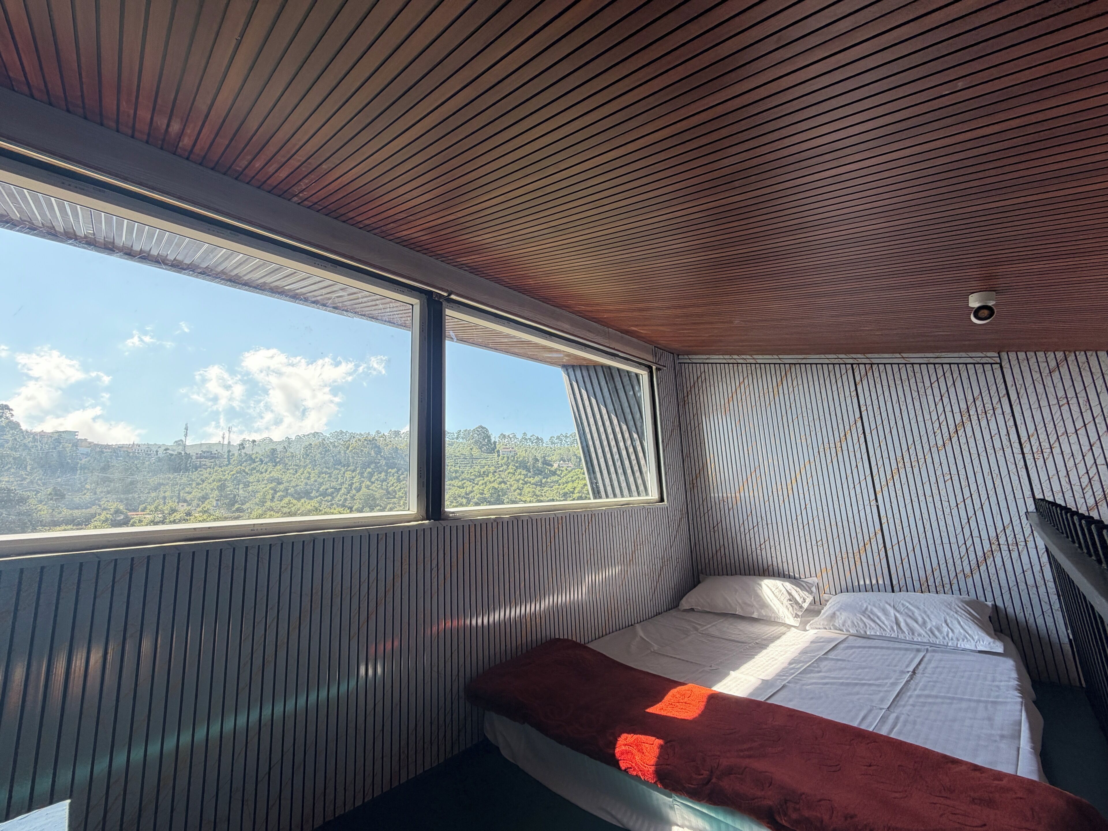 A room with a bed, a window, and a view of a forested area.