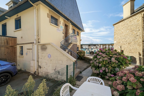 Un bâtiment avec un escalier menant à un balcon, une voiture garée et un jardin agrémenté d’hortensias.