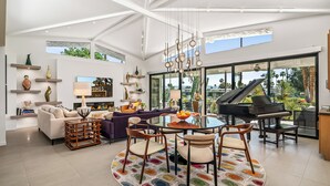 Een ruime woonkamer met een piano, een ronde eettafel en een groot raam met uitzicht op een tuin.