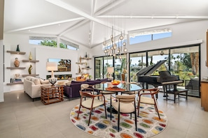 Ein geräumiges Wohnzimmer mit einem Klavier, einem runden Esstisch und einem großen Fenster mit Blick auf einen Garten.