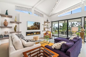 A modern living room with a fireplace, a large window with a view of palm trees, and a mounted artwork of palm trees. - The Fairway Flat: MCM Gem with Golf, Tennis, Pickleball, Fitness Center (Palm Springs)