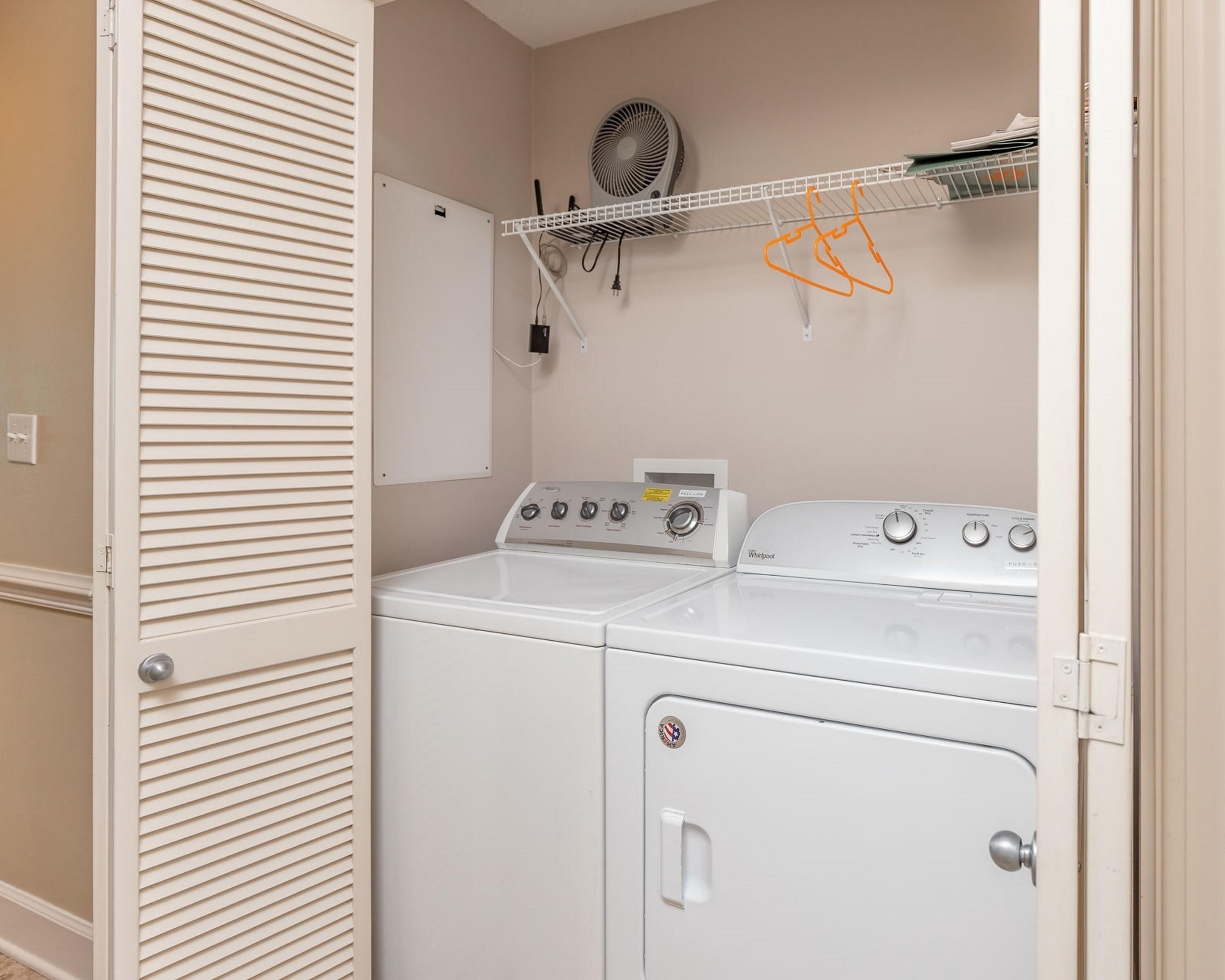 A laundry room with two washers and dryers, a fan, and a shelf with items.