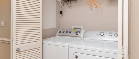 A laundry room with two washers and dryers, a fan, and a shelf with items.