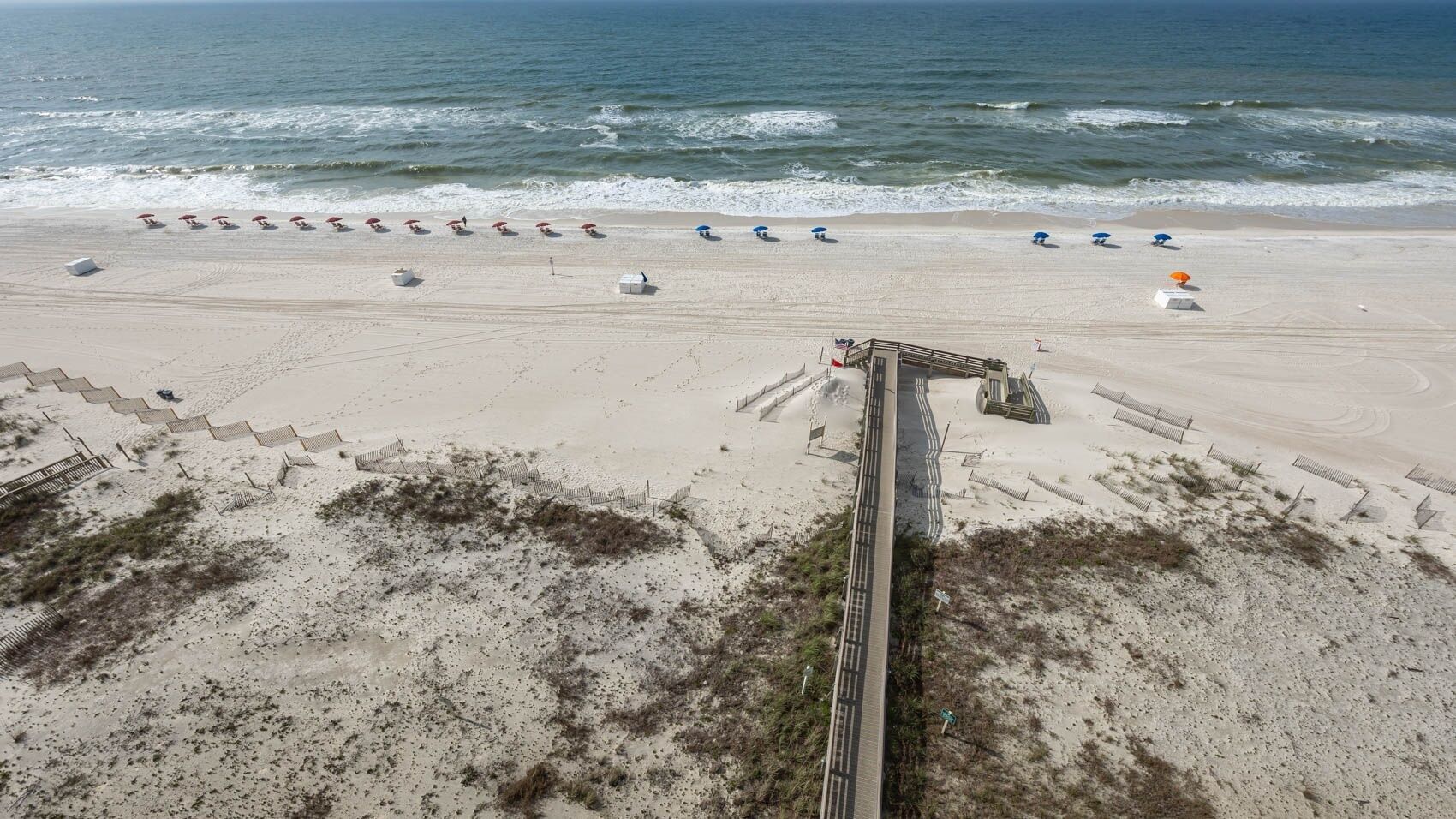 A beach with a long wooden boardwalk leading to a lifeguard tower. There are several beach umbrellas and lounge chairs along the shore.
