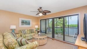 A living room with a ceiling fan, a glass coffee table, and a sofa with a tropical print. - First-floor, snowbird-friendly townhouse w/ shared pool, WiFi, central AC, & W/D (Bokeelia)