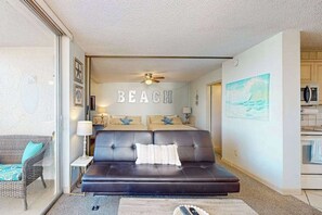 A room with a dark leather sofa, a glass door leading to a balcony, a ceiling fan, and a wall with the word 'BEACH' spelled out. - Renovated Beachfront Condo: Modern Comfort, River Views & Endless Relaxation! (Bonita Springs)