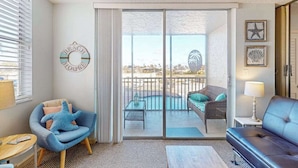 A room with a sliding glass door leading to a balcony with a pool, a blue armchair, a sofa, and a small table. - Renovated Beachfront Condo: Modern Comfort, River Views & Endless Relaxation! (Bonita Springs)