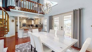 Uma ampla área de refeições com uma grande mesa de mármore e cadeiras brancas, uma cozinha ao fundo e uma escada que leva a um andar superior.