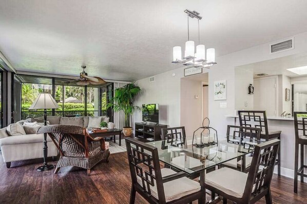 A modern living room with a glass dining table, dark wooden chairs, a wicker sofa, and a ceiling fan. - Spacious, air-conditioned condo with private W/D, beach shuttle, & central AC (Bonita Springs)