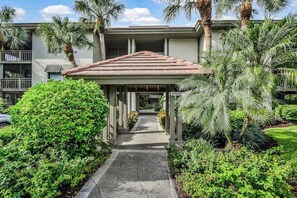 A covered walkway leading to a building with a tiled roof, surrounded by lush greenery and palm trees. - Spacious, air-conditioned condo with private W/D, beach shuttle, & central AC (Bonita Springs)