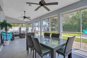 Une vaste terrasse dotée d’une table en verre et de chaises en osier, de ventilateurs de plafond et de grandes fenêtres offrant une vue sur l’extérieur.