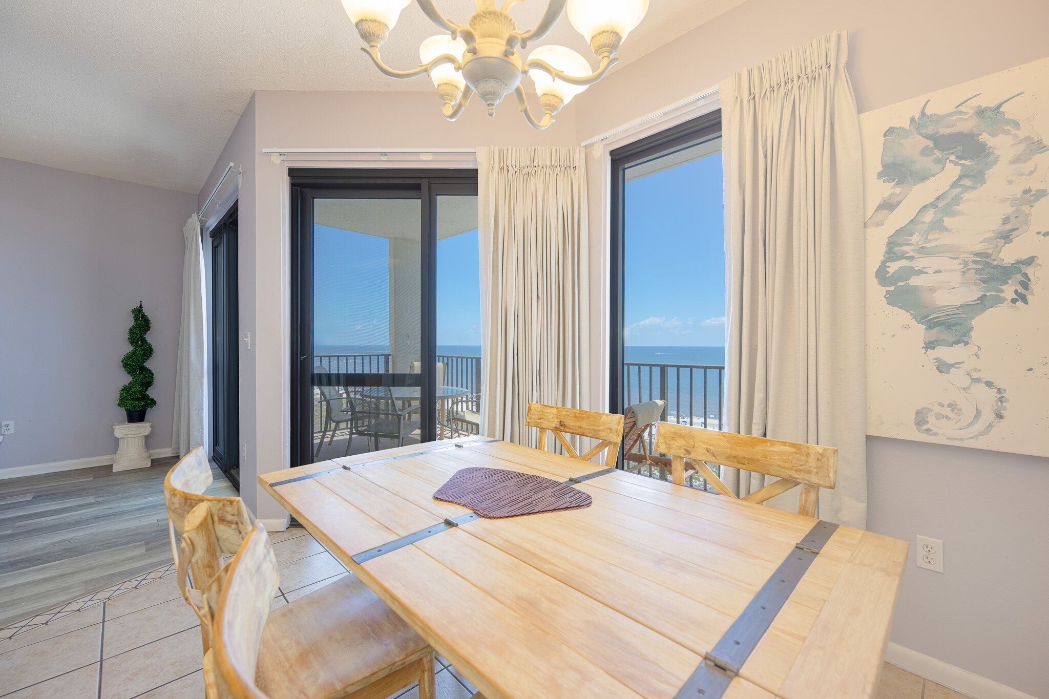 A dining area with a wooden table and chairs, a chandelier, and a view of the sea through large windows.