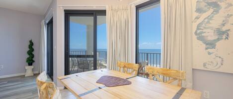 A dining area with a wooden table and chairs, a chandelier, and a view of the sea through large windows.
