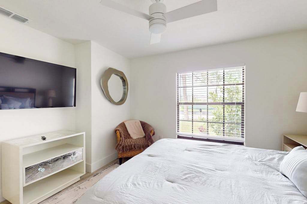 A bedroom with a bed, a TV mounted on the wall, a window with blinds, a chair, and a ceiling fan.