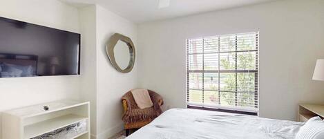 A bedroom with a bed, a TV mounted on the wall, a window with blinds, a chair, and a ceiling fan.