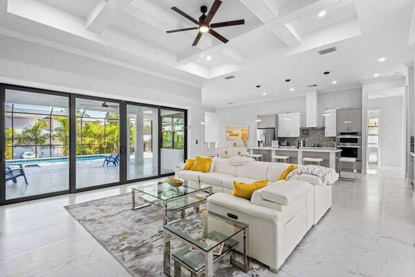A spacious living area with a glass coffee table, a white sofa with yellow cushions, and a ceiling fan. - Luxe brand-new home with designer interior, lanai, private pool, & spa (Bonita Springs)