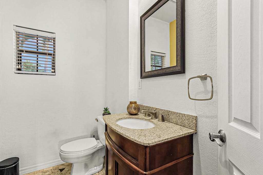 Un baño con inodoro blanco, lavamanos con tocador de madera, espejo y ventana con persianas.