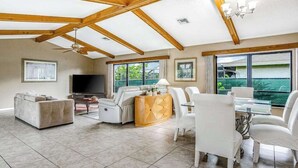 A spacious living room with a high ceiling, built-in wooden beams, a flat-screen TV, a sofa, a dining table with chairs, and large windows offering a view. - Spacious waterfront home with private pool & grill - near downtown (Cape Coral)