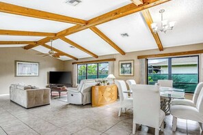 A spacious living room with a high ceiling, built-in wooden beams, a flat-screen TV, a sofa, a dining table with chairs, and large windows offering a view. - Spacious waterfront home with private pool & grill - near downtown (Cape Coral)