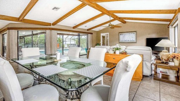 A spacious room with a glass dining table, white chairs, a sofa, a TV, and a wooden ceiling structure. - Spacious waterfront home with private pool & grill - near downtown (Cape Coral)