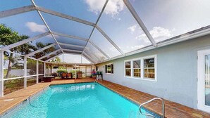 Una piscina con cerramiento de vidrio, un patio y una casa con fachada azul claro.