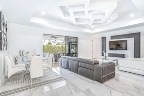 A modern living room with a sectional sofa, a flat-screen TV, and a dining area with white furniture. - Dog-friendly pool home with outdoor kitchen, game room, cabana, and pool spa (Cape Coral)