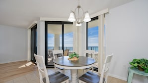 A dining area with a round table, white chairs, a chandelier, and a view through a glass door. - Phoenix Vi 6607 1 Bedroom Condo (Orange Beach)